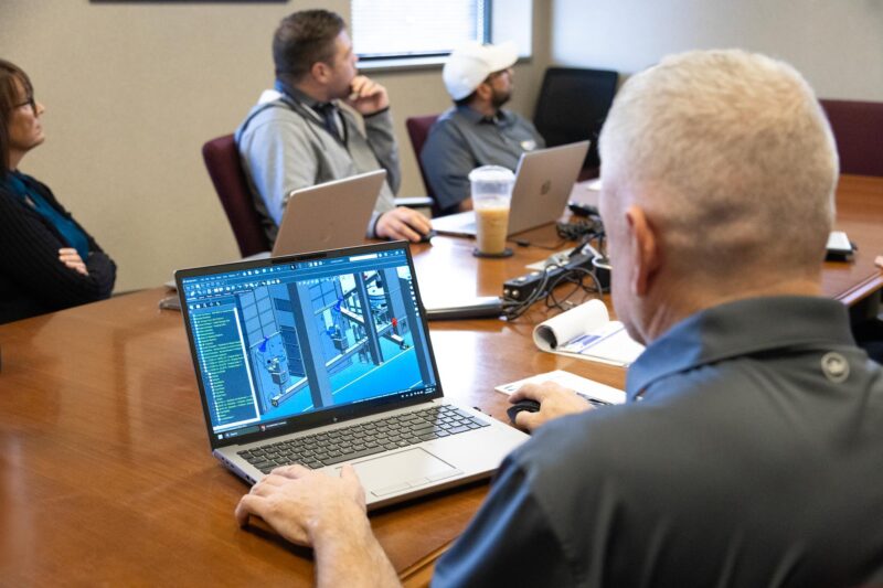 consulting services; people around a conference table with a laptop computer showing factory machine renderings